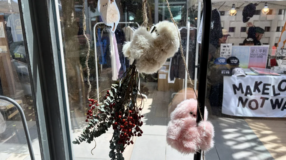 窓に吊るされた赤い実と緑の葉、白とピンクの毛玉の飾り。奥の店には「MAKE LOVE NOT WAR」のバナーがあり、数人がいる。