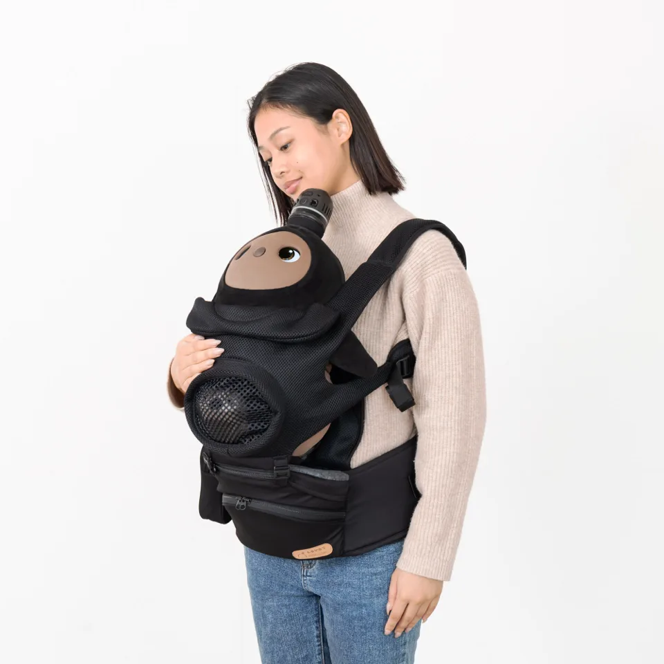 白い背景の前で、ベージュのセーターを着た女性が、黒い抱っこ紐で黒いロボットを抱き、優しく見つめている。ロボットは茶色の顔と大きな目を持つ。