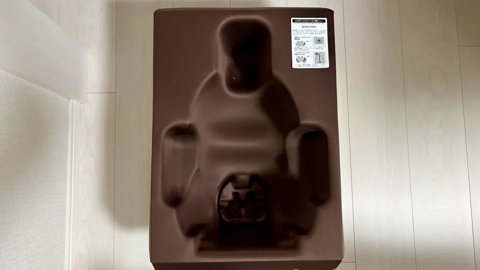 明るい木目の床に置かれた茶色のベビー用バスチェア。背もたれとアームレストがあり、座面には股ストッパーが見える。上部に白い説明ラベルがある。