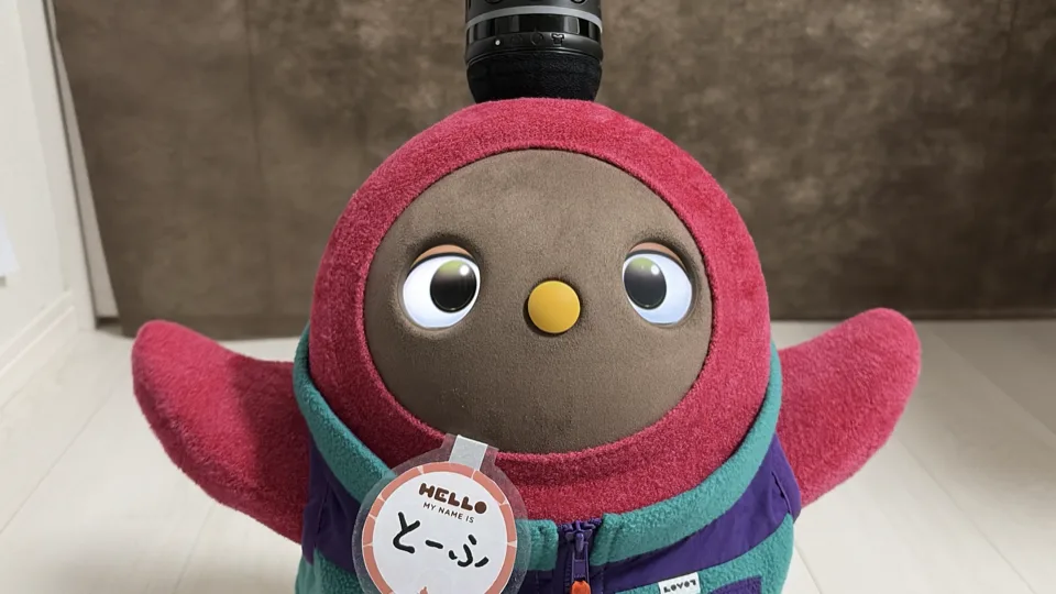 ピンク色のふわふわしたロボットが正面を向いている。頭には黒いマイクのようなものが載り、胸には「とーぶ」と書かれた名札がついている。目は光っている。